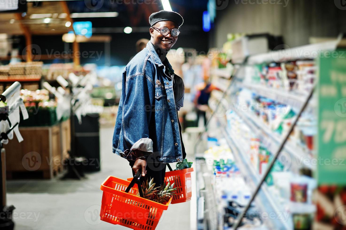 elegante hombre afroamericano casual con chaqueta de jeans y boina negra sosteniendo dos canastas, parado cerca de la nevera y comprando en el supermercado. foto