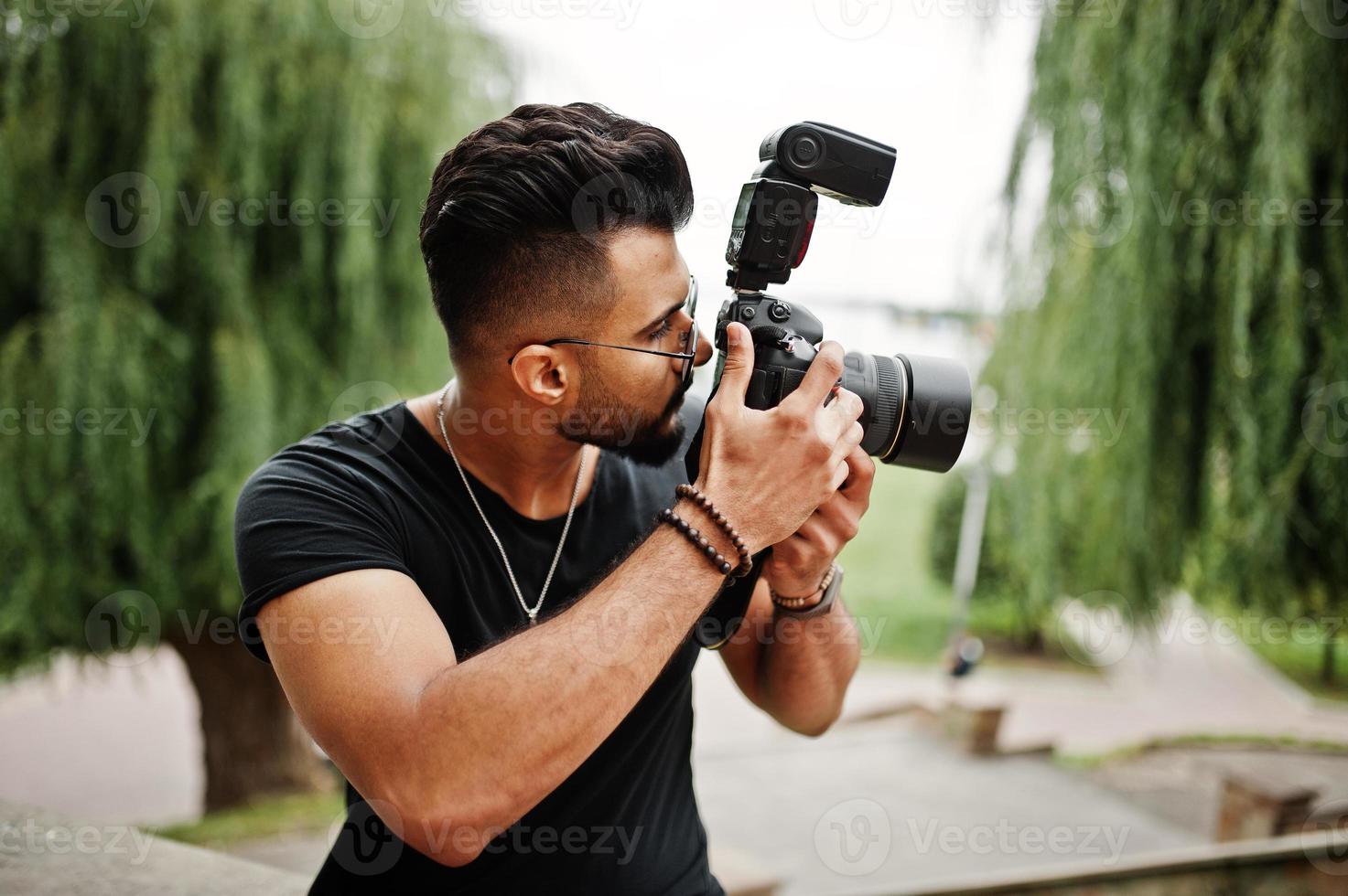 Awesome beautiful tall ararbian beard macho man photographer in glasses and black t-shirt with professional camera at hands. photo