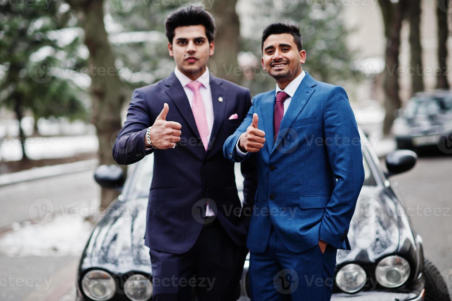Two elegant indian fashionable mans model on suit posed at winter day against black classic car and shows thumbs up. photo