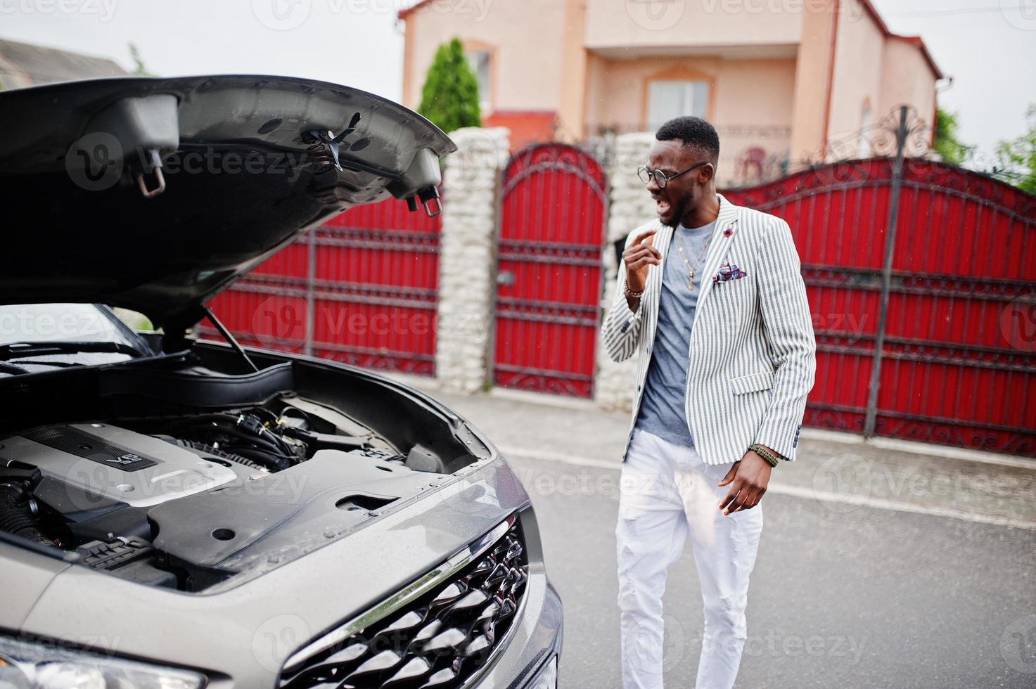 Stylish and rich african american man stand in front of a broken suv car needs assistance looking under opened hood. photo