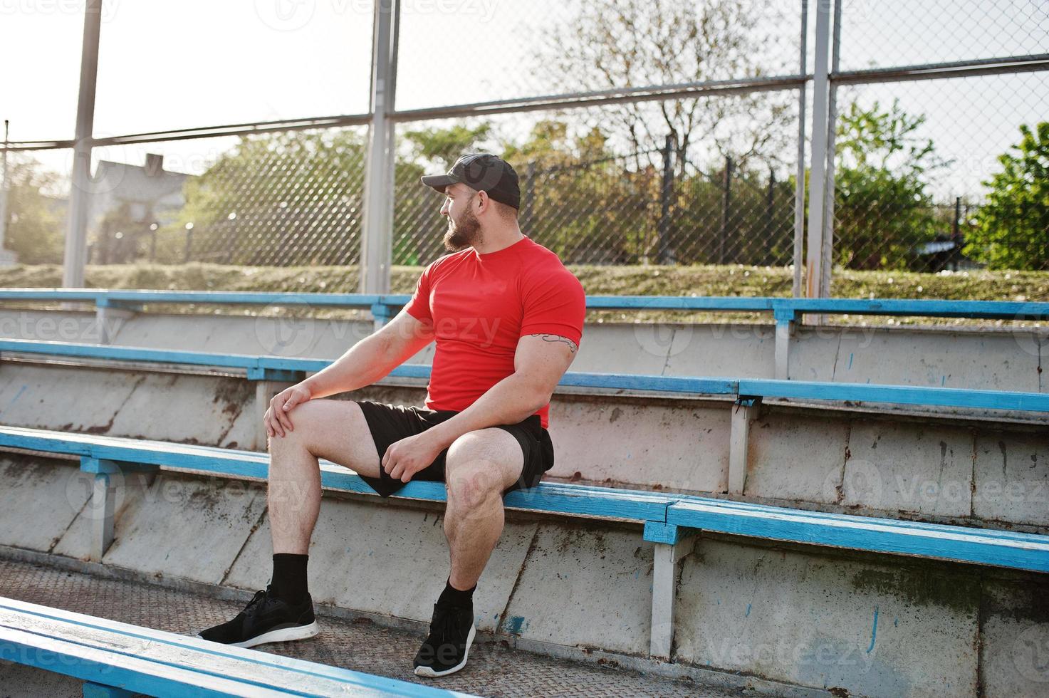 Young brutal bearded muscular man wear on red shirt, shorts and cap at stadium. photo