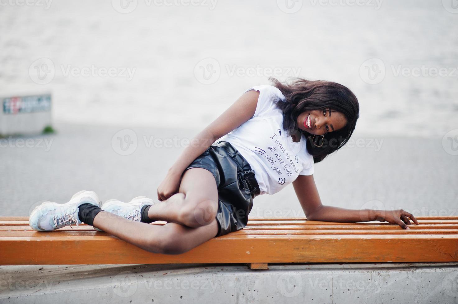 Modelo delgado afroamericano de piel oscura posado en pantalones cortos de cuero negro y camiseta blanca. ella luying en banco. foto