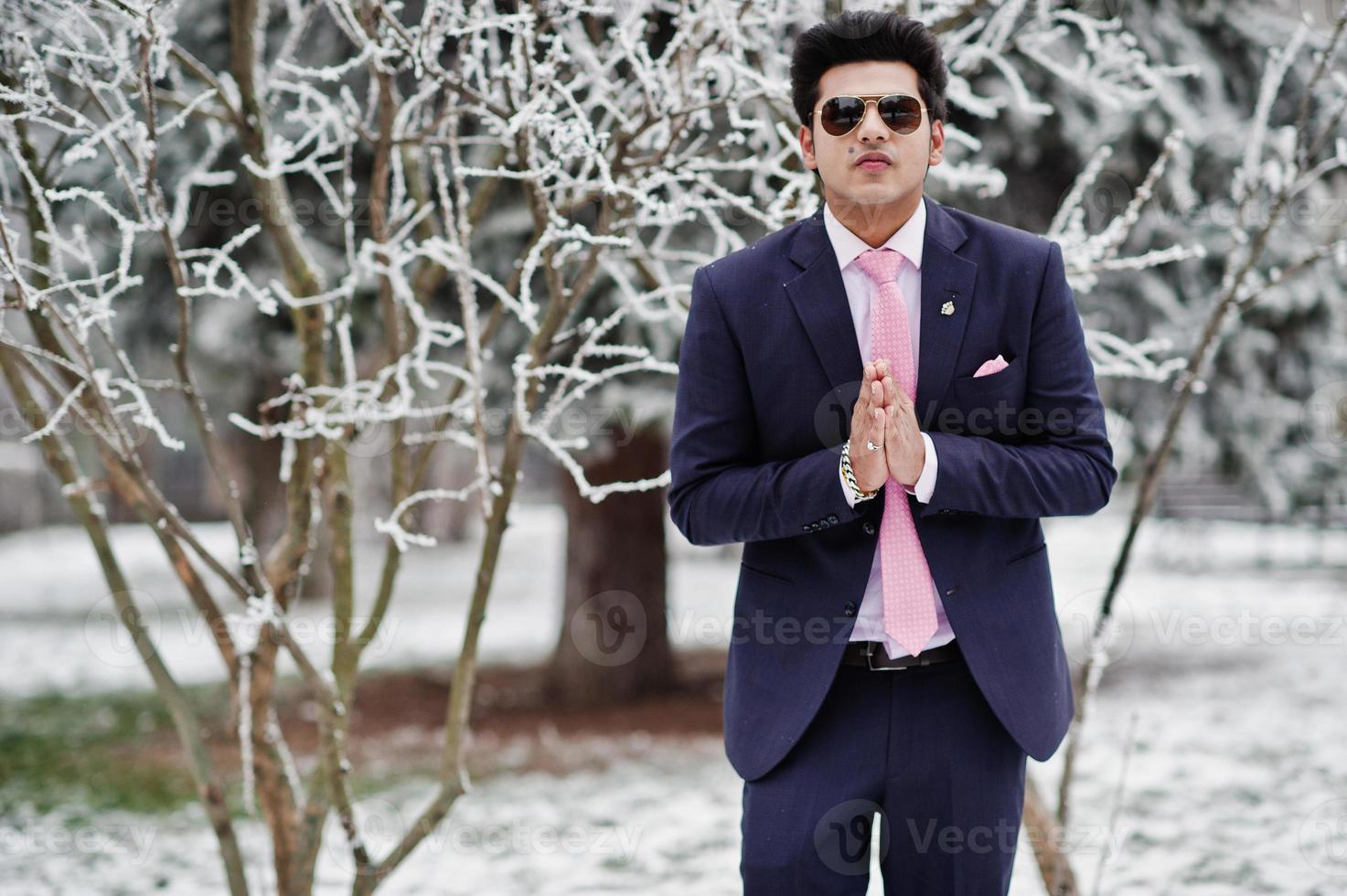 Elegant indian macho man model on suit and pink tie, sunglasses posed on winter day shows namaste. photo
