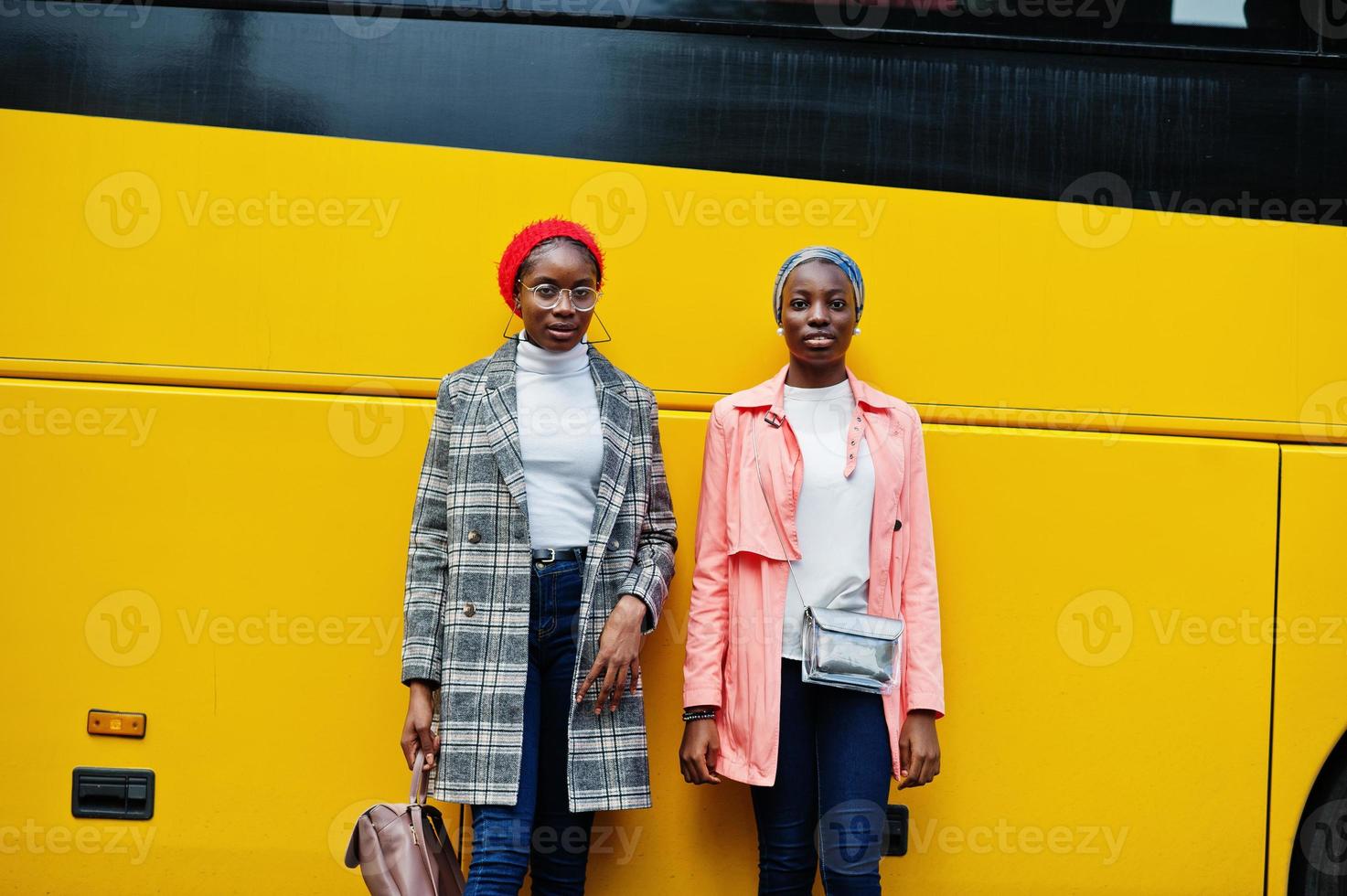 Two young modern fashionable, attractive, tall and slim african muslim womans in hijab or turban head scarf and coat posed against yellow bus. photo
