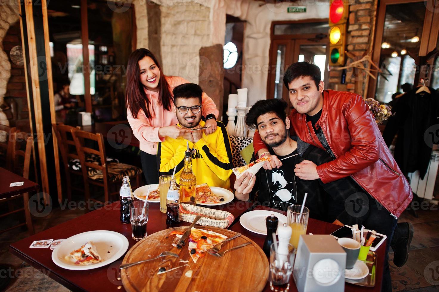 Group of asian friends eating pizza during party at pizzeria. Happy indian people having fun together, eating italian food and sitting on couch, fighting with forks and knifes. photo