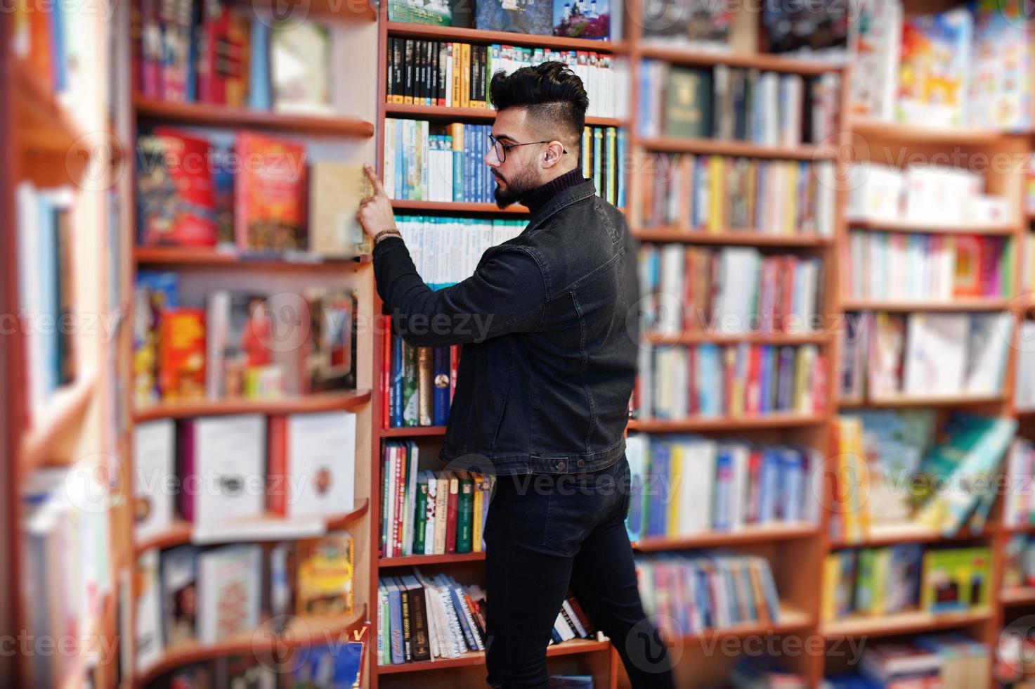 estudiante árabe alto e inteligente, vestido con chaqueta de jeans negros y anteojos, en la biblioteca seleccionando un libro. foto