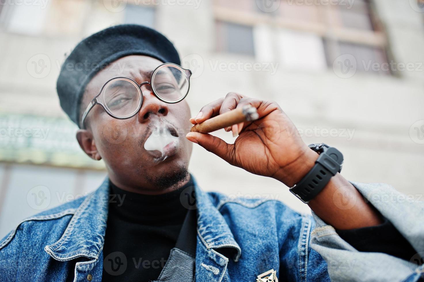 Close up portrait of african american man in jeans jacket, beret and eyeglasses, smoking cigar. photo