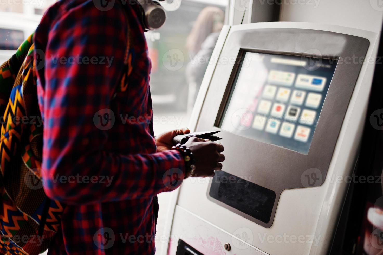 African american man in checkered shirt with backpack holding wallet in hand against atm. Black man traveler. photo