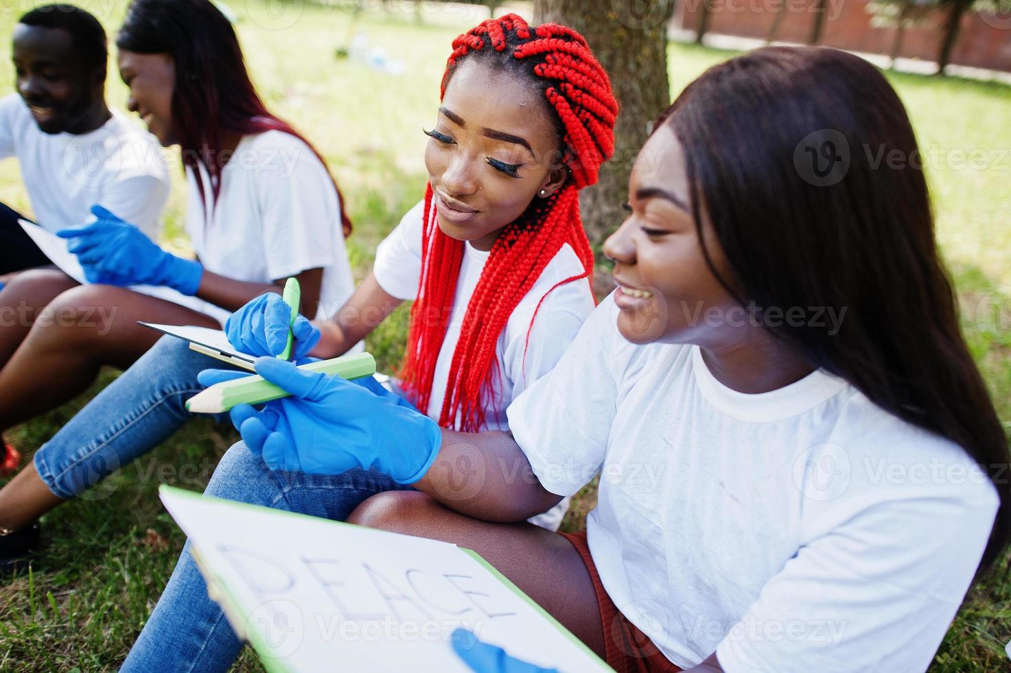 Group of happy african volunteers sitting under tree at park and write something on clipboards. Africa volunteering, charity, people and ecology concept. photo