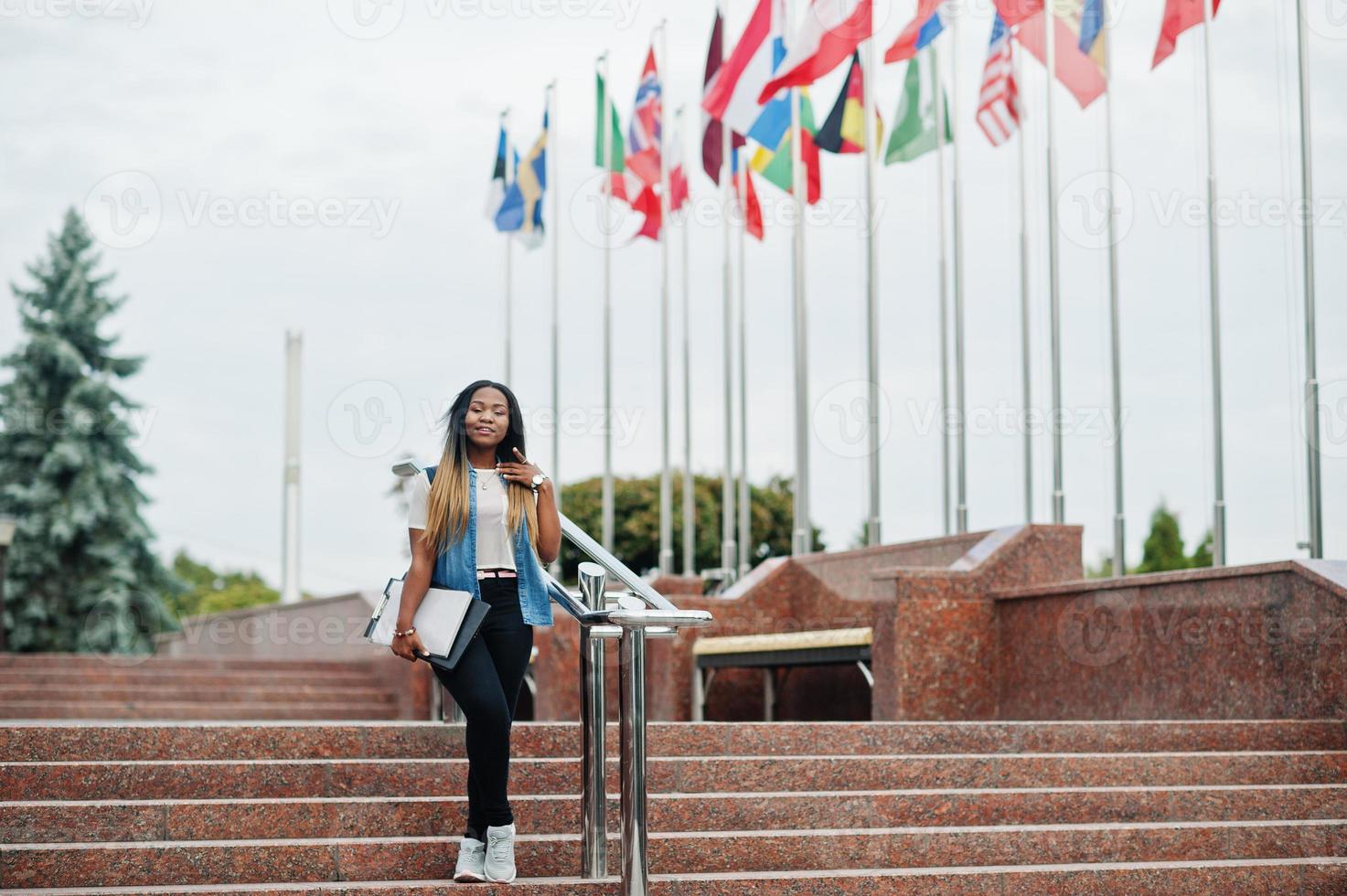 African student female posed with backpack and school items on yard of university, against flags of different countries. photo