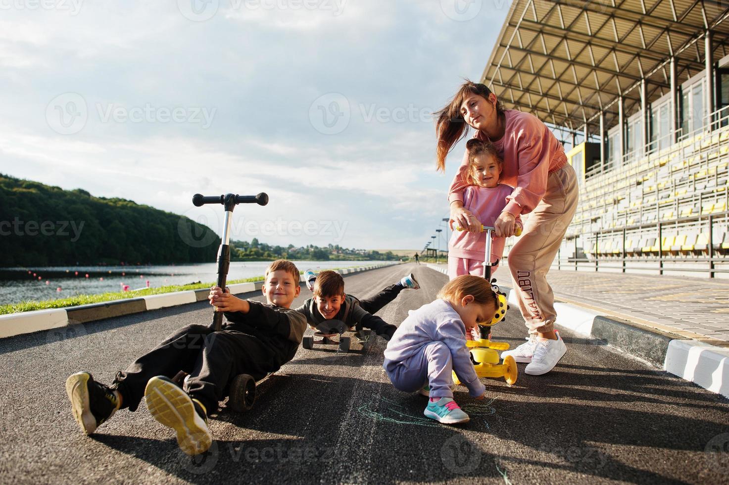 Young stylish mother with four kids outdoor. Sports family spend free time outdoors with scooters and skates. photo