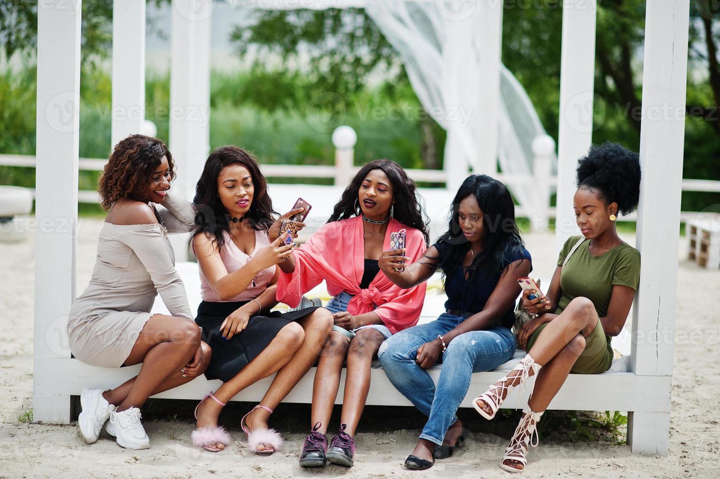 Group of five african american girls relaxing at beautiful poolside cabana beside luxury resort and looking at mobile phones. photo