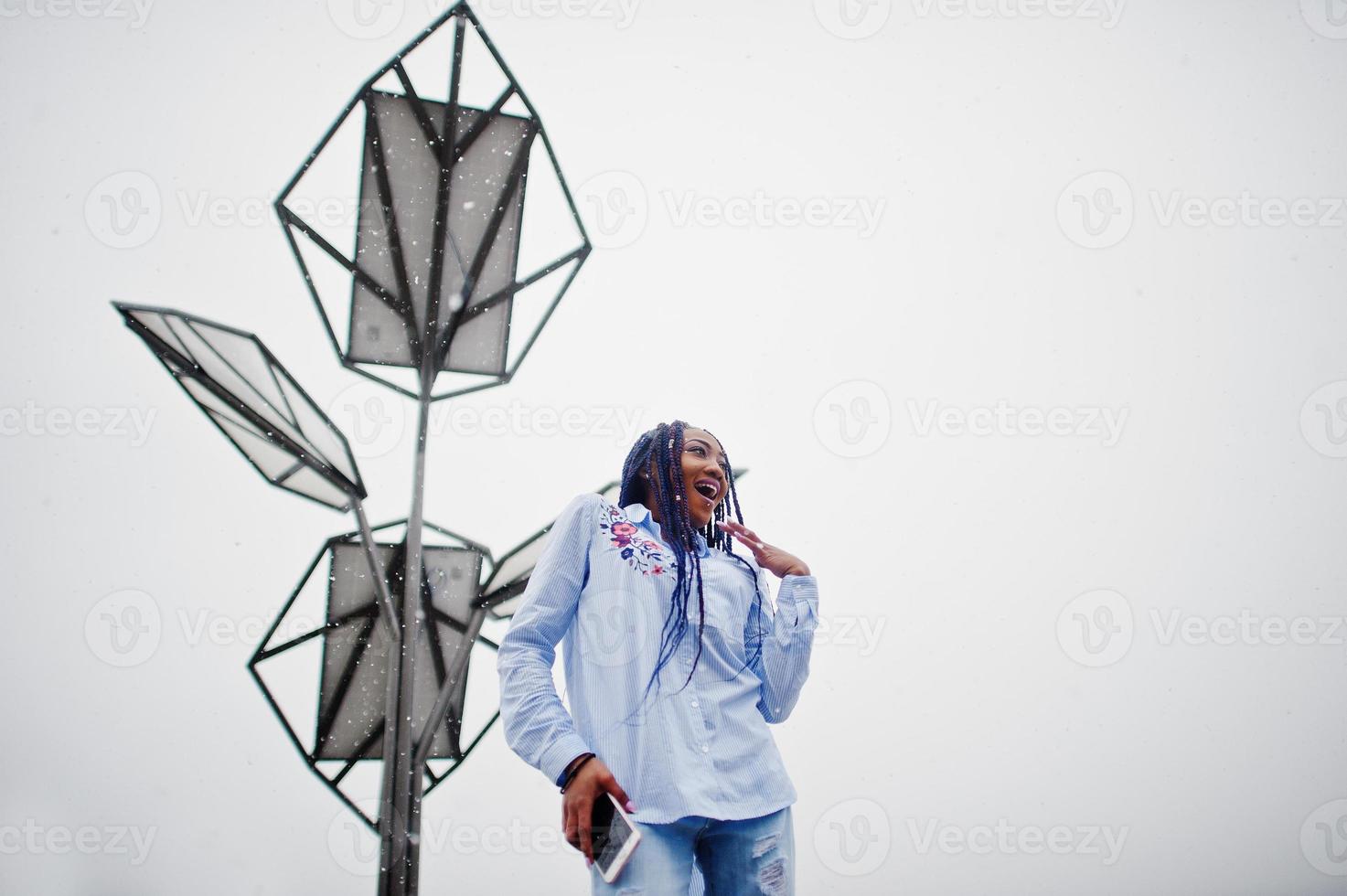 Stylish african american girl with dreads holding mobile phone at hand, outdoor against solar batteries at snowy weather. photo