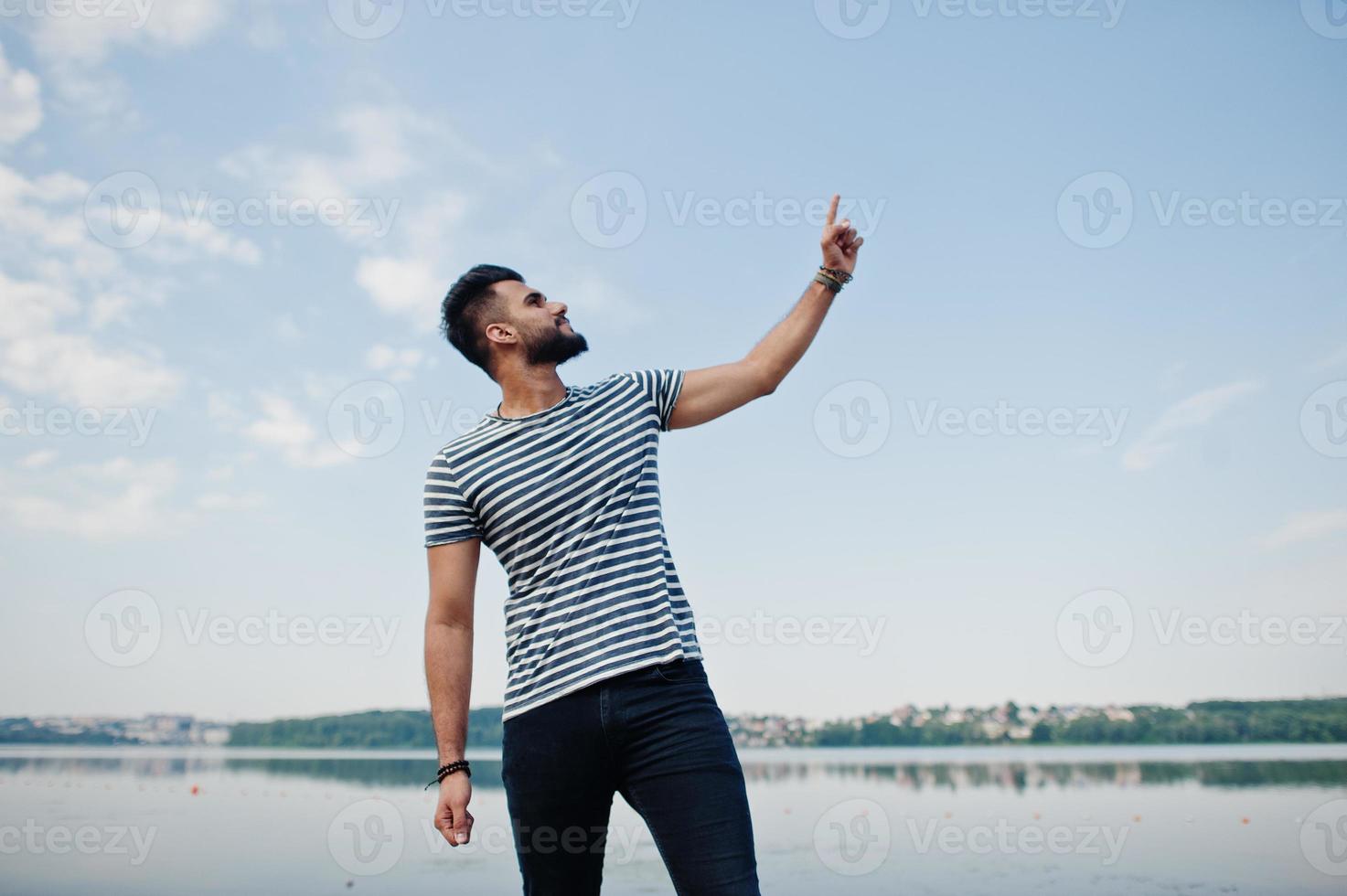 modelo de hombre de barba árabe alto y guapo en camisa desnuda posada al aire libre contra el lago y mostrando el dedo hacia el cielo. chico árabe de moda. foto