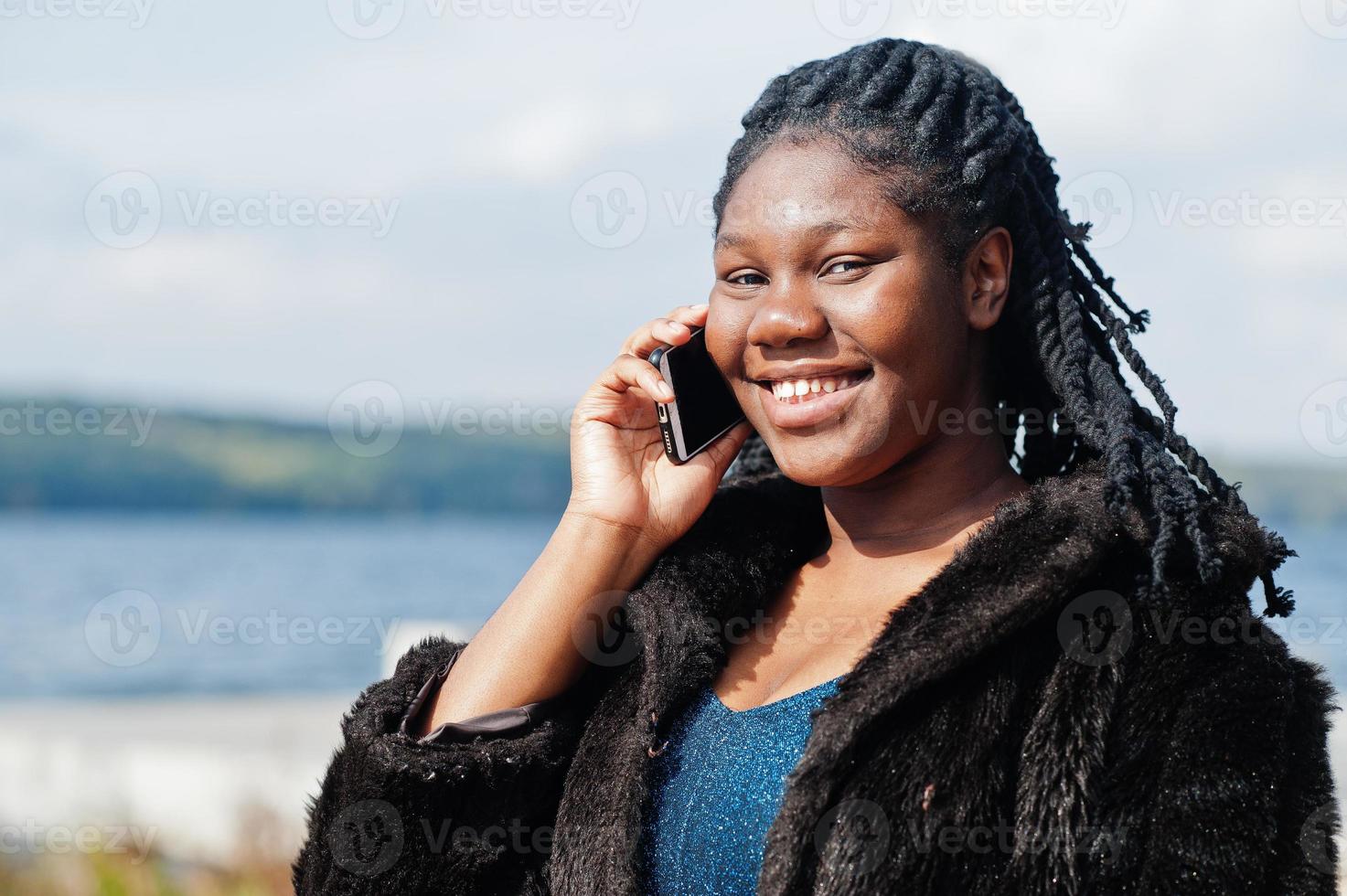 modelo afroamericano de piel oscura de talla grande posado en un vestido  azul brillante y abrigo de piel negro contra el mar habla por teléfono.  10501465 Foto de stock en Vecteezy