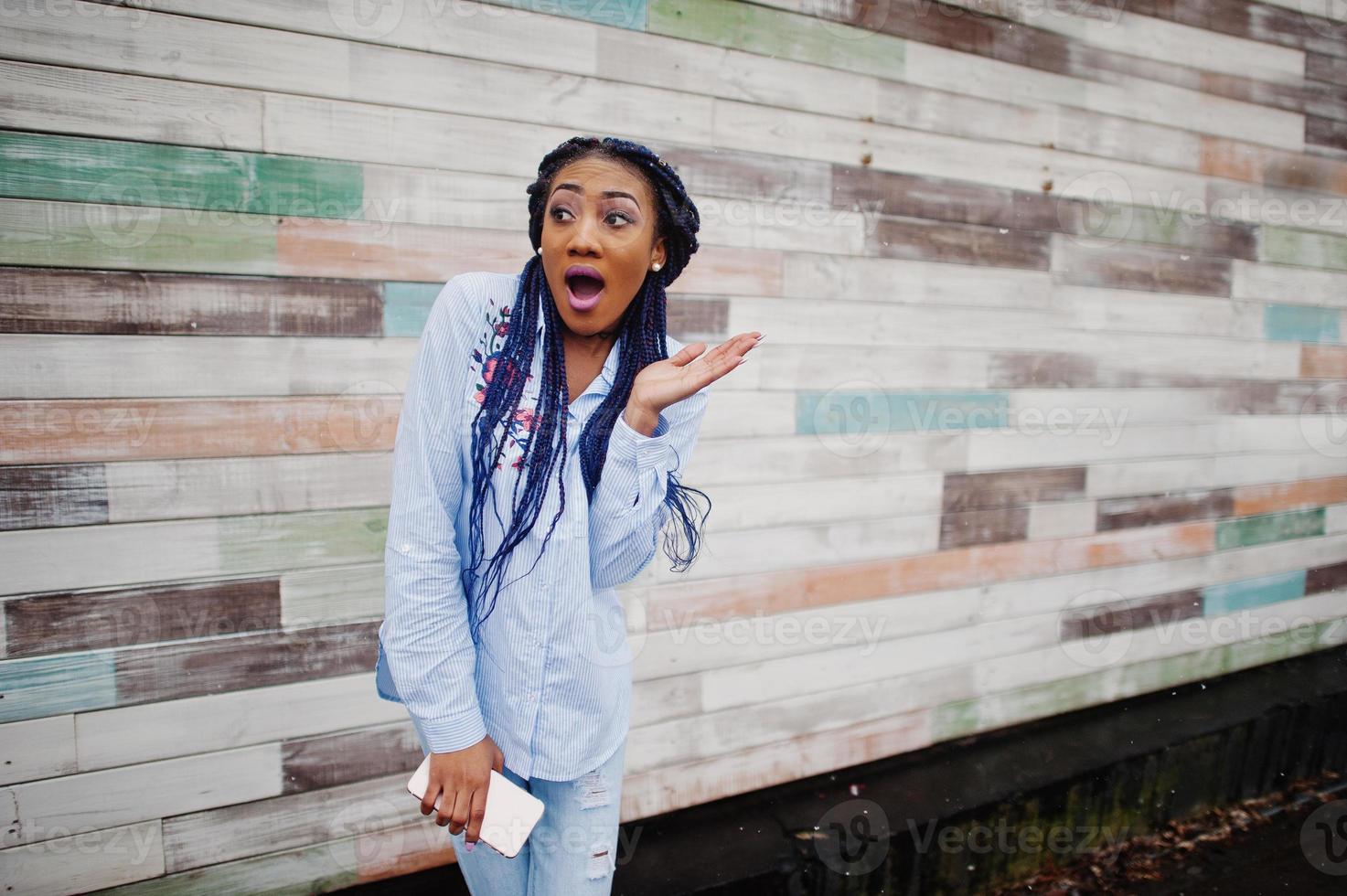 Stylish african american girl with dreads holding mobile phone at hand, outdoor against wooden wall. photo
