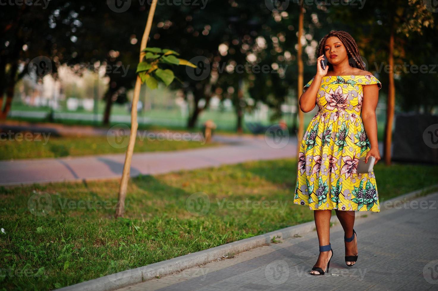Cute small height african american girl with dreadlocks, wear at coloured yellow dress, posed at sunset, walking and speaking on cellphone. photo