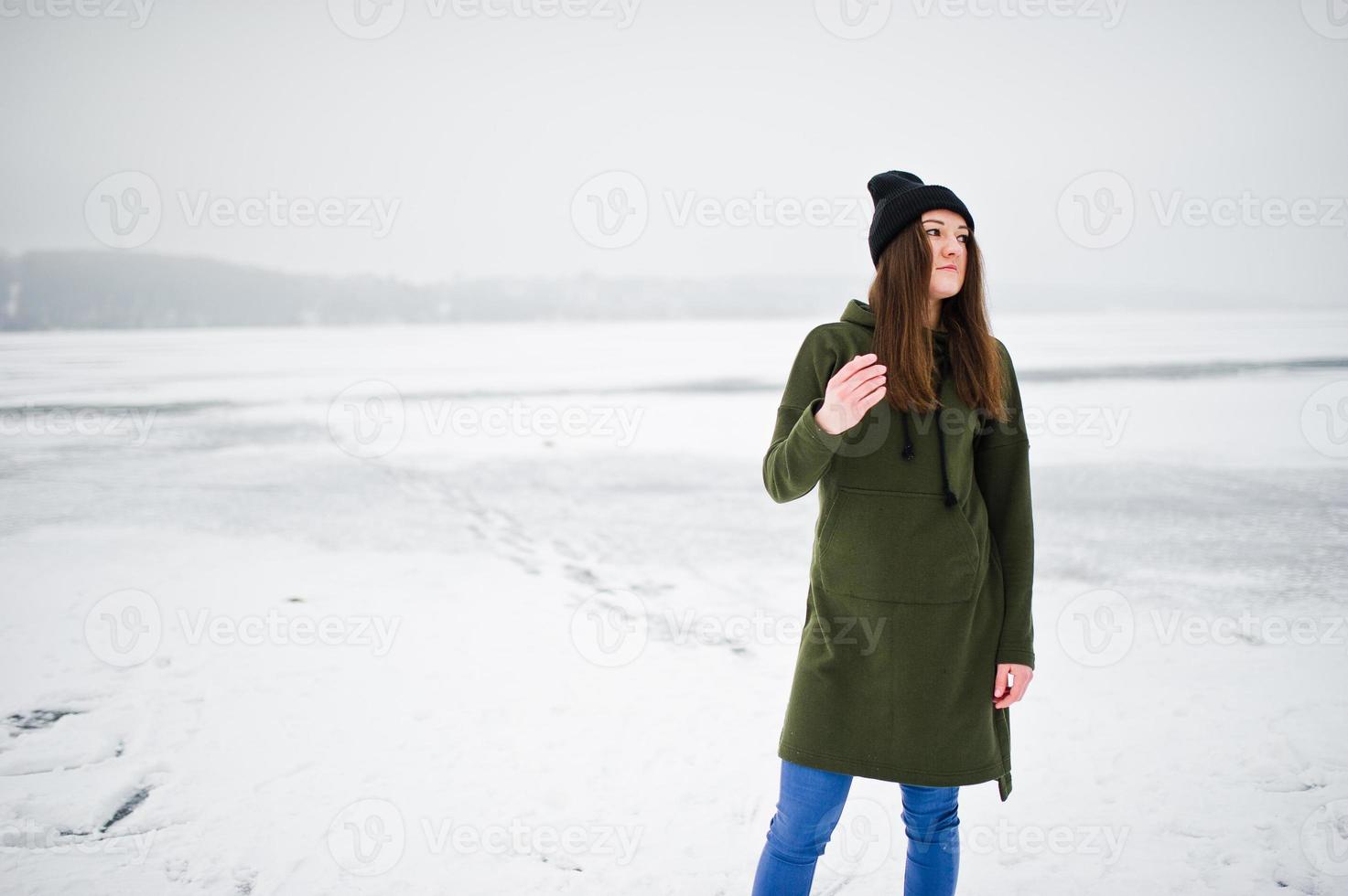 Funny girl wear on long green sweatshirt, jeans and black headwear, at frozen lake in winter day. photo