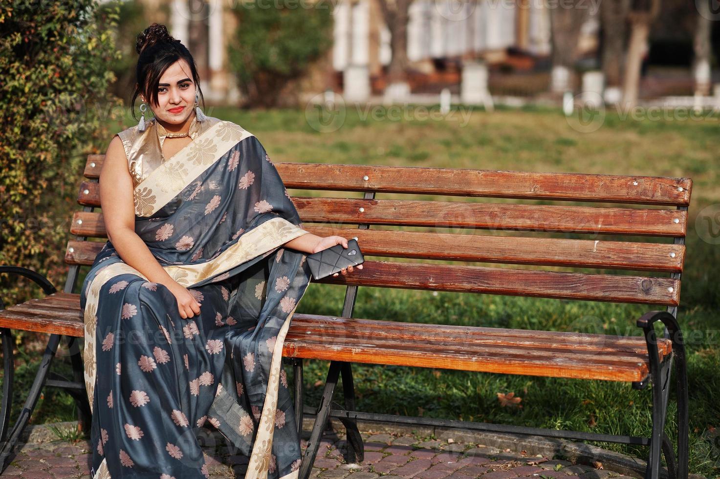 Elegant brunette south asian indian girl in saree sitting on bench with mobile phone at hands. photo