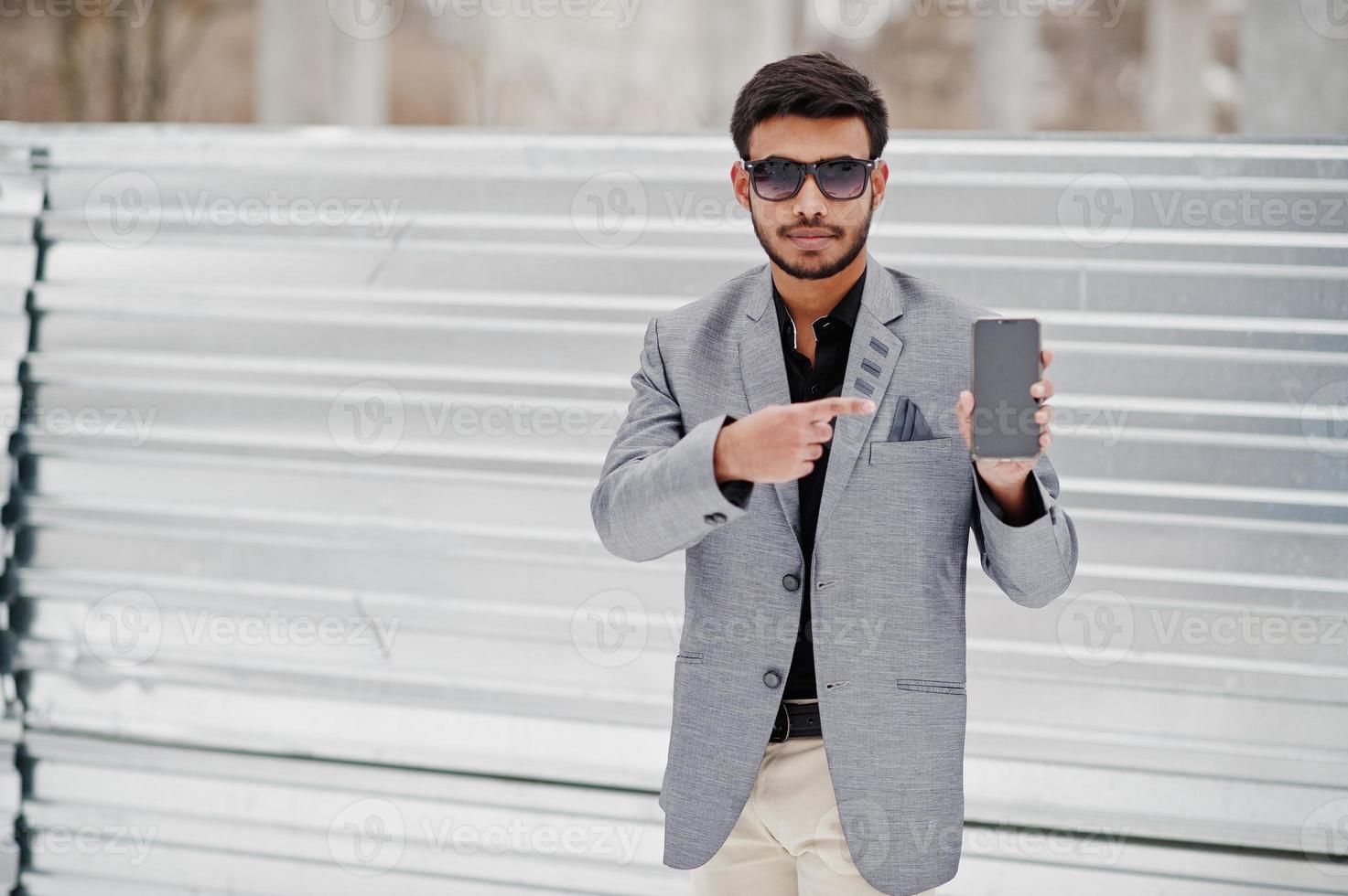 Casual young indian man in silver jacket and sunglasses posed against steel background, showing to mobile phone. photo