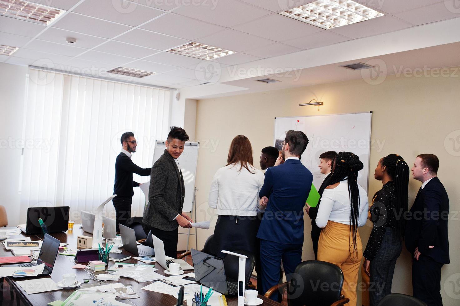 Mixed race business coach presenting report standing near whiteboard pointing on sales statistic shown on diagram and chart teach diverse company members gathered together in conference room. photo