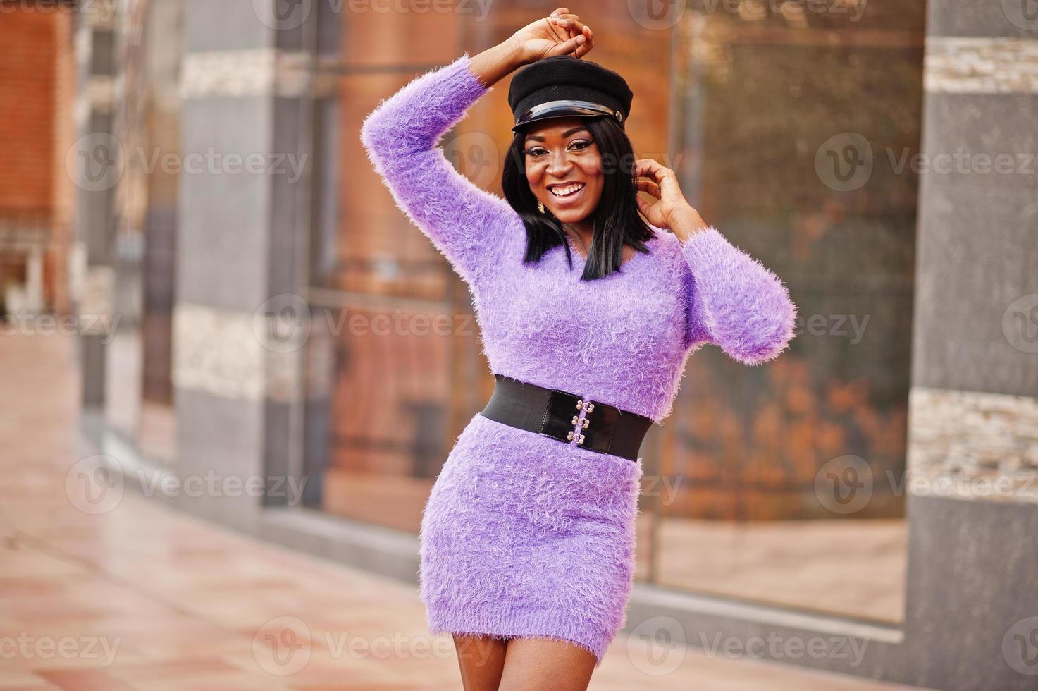 African american woman at violet dress and cap posed outdoor against modern building. photo