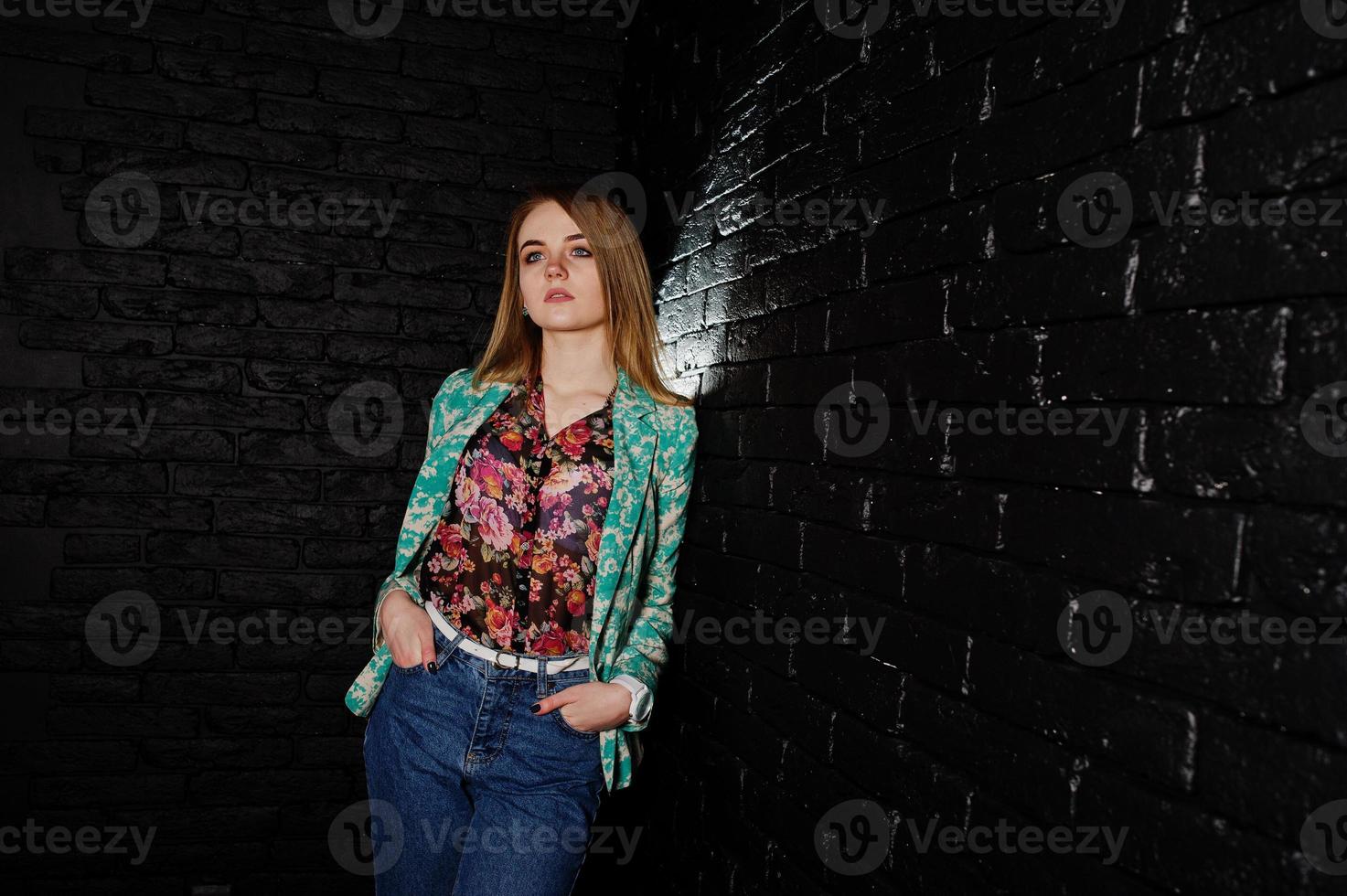 chica rubia con estilo en chaqueta y jeans contra la pared de ladrillo negro en el estudio. foto