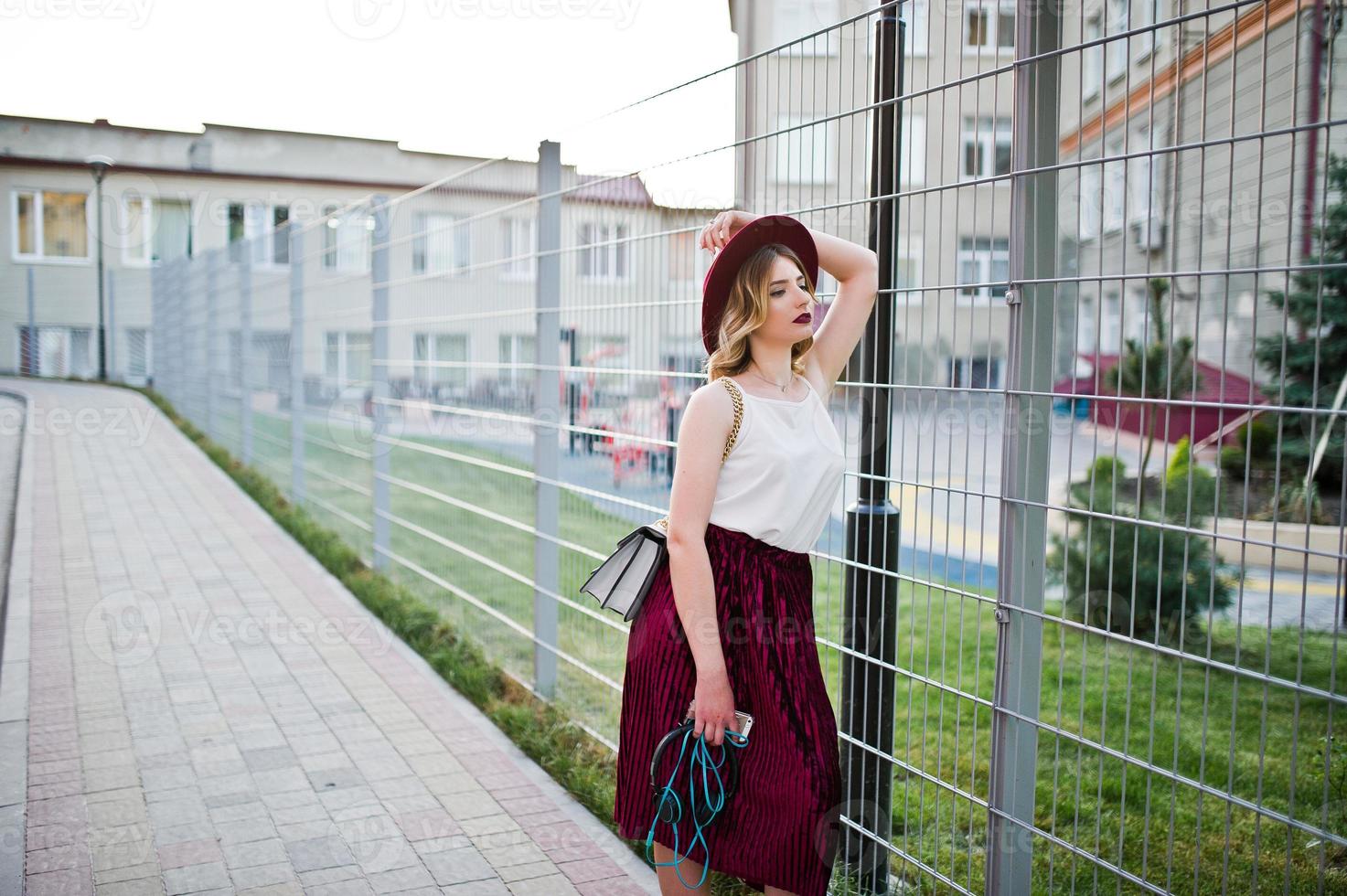 Fashionable and beautiful blonde model girl in stylish red velvet velour skirt, white blouse and hat, posed with phone and earphones. photo