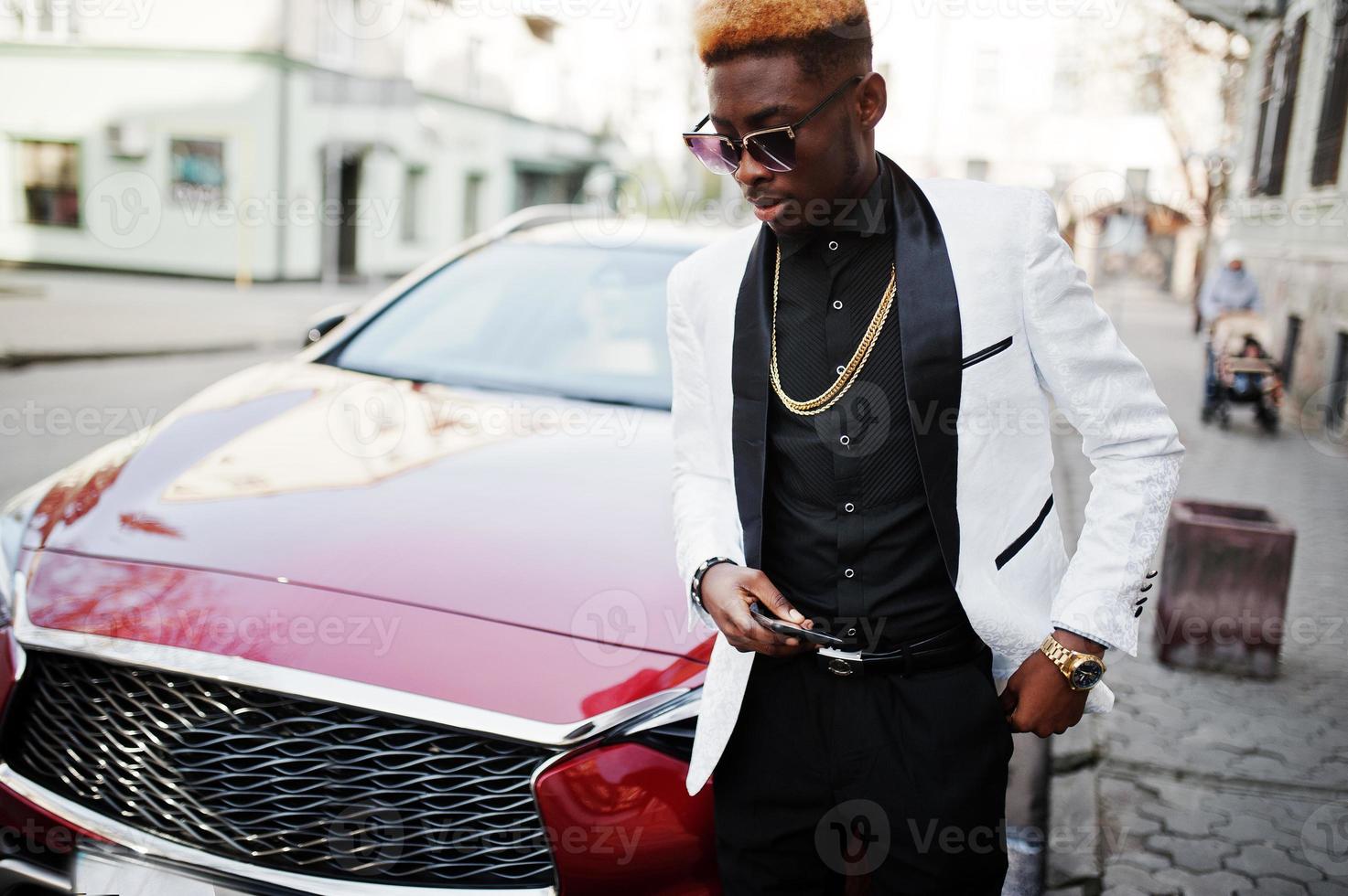 hombre afroamericano elegante y apuesto con traje blanco contra un auto rojo de lujo con teléfono móvil a mano. foto