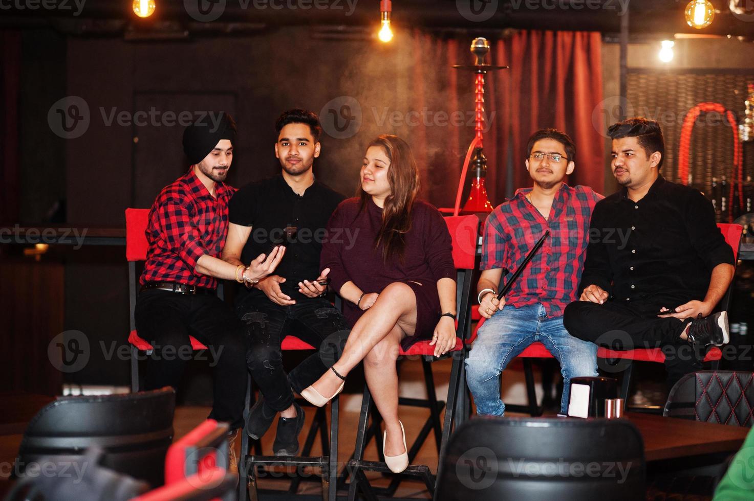 Group of indian friends sitting at lounge bar, having fun and rest, smoking hookah and looking on mobile phones. photo