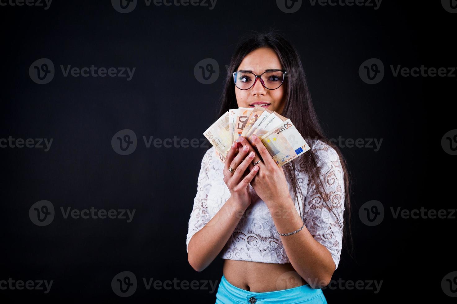 retrato de una chica atractiva con camiseta azul o turquesa, pantalones y gafas posando con mucho dinero en la mano. foto