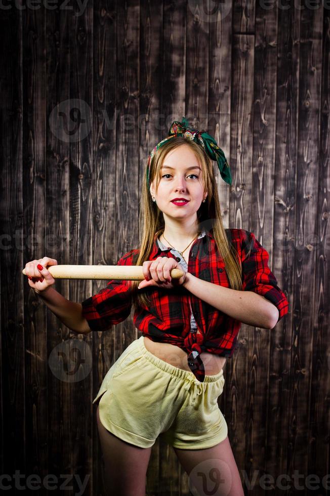 joven ama de casa divertida en camisa a cuadros y pantalones cortos amarillos estilo pin up con rodillo de cocina sobre fondo de madera. foto