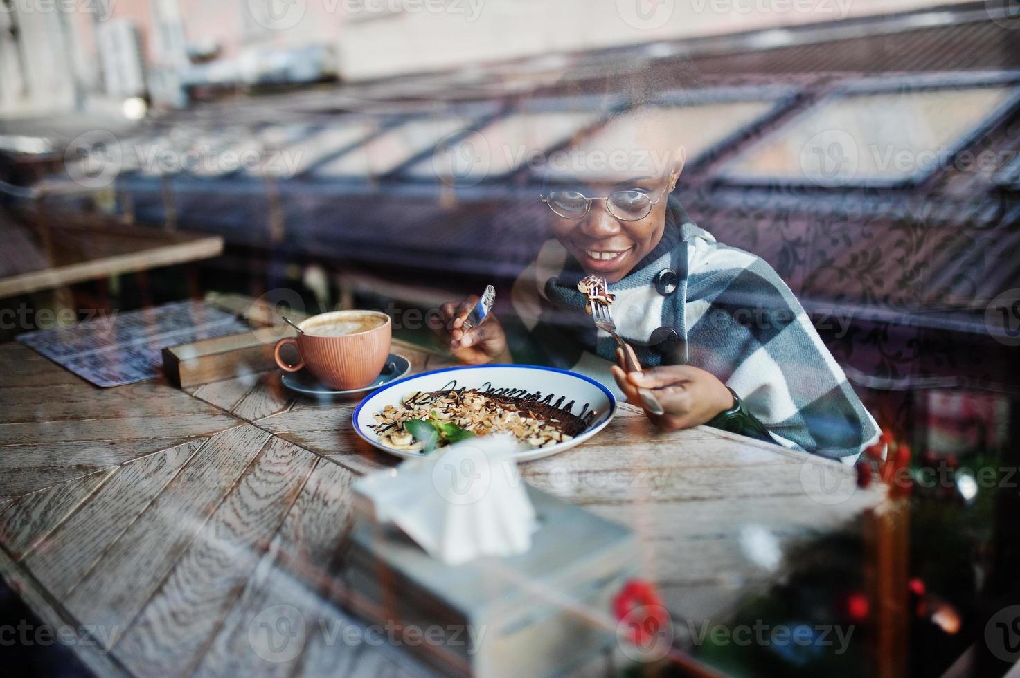 African woman in checkered cape and eyeglasses sitting at cafe and eating dessert. photo