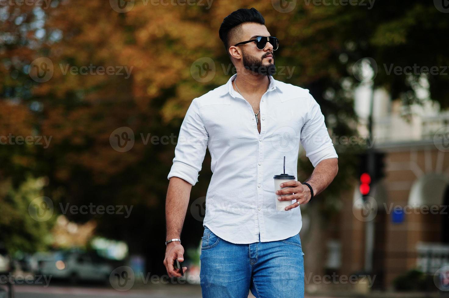 Stylish tall arabian man model in white shirt, jeans and sunglasses posed at street of city. Beard attractive arab guy with cup of coffee walking. photo