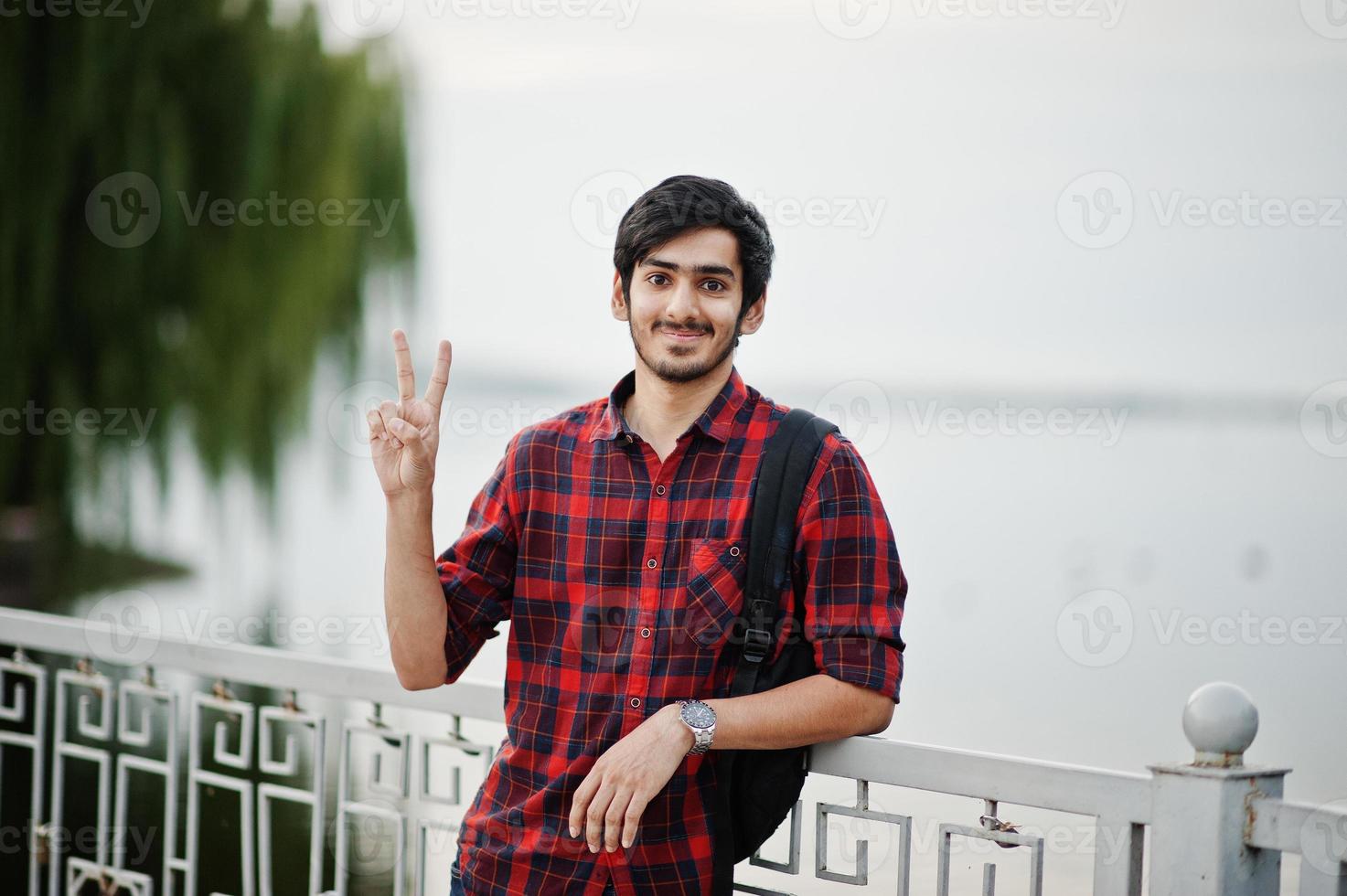 Young indian student man at checkered shirt and jeans with backpack posed on evening city against lake, shows two fingers. photo