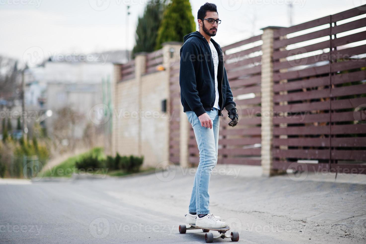 Street style arab man in eyeglasses with longboard longboarding down the road. photo