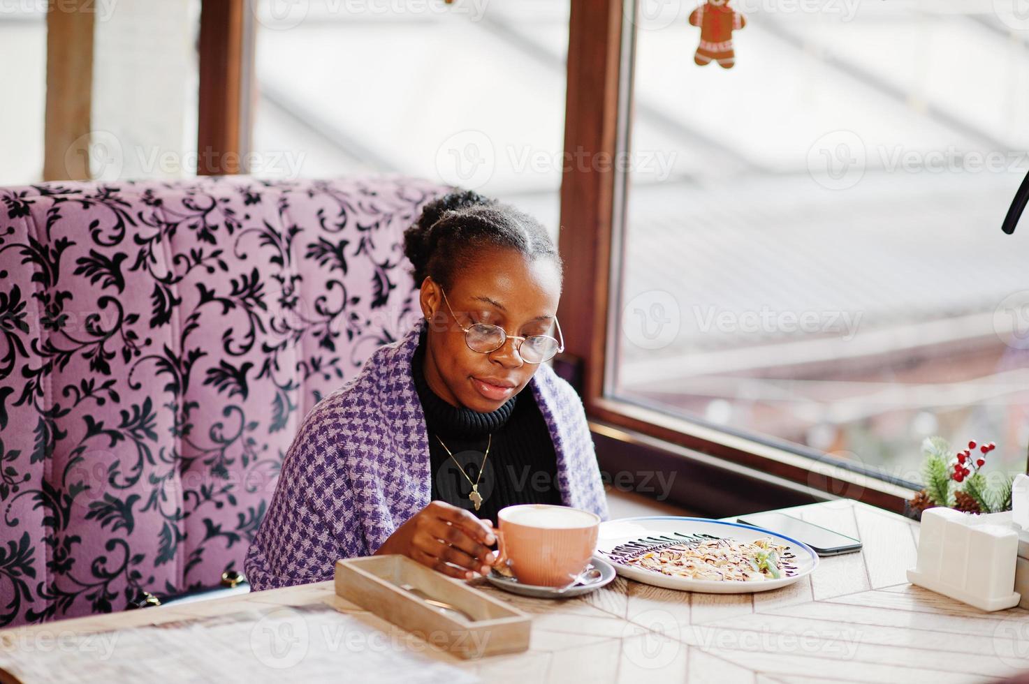 African woman in checkered violet cape and eyeglasses posed at cafe, sitting by table with dessert and cup of coffee. photo