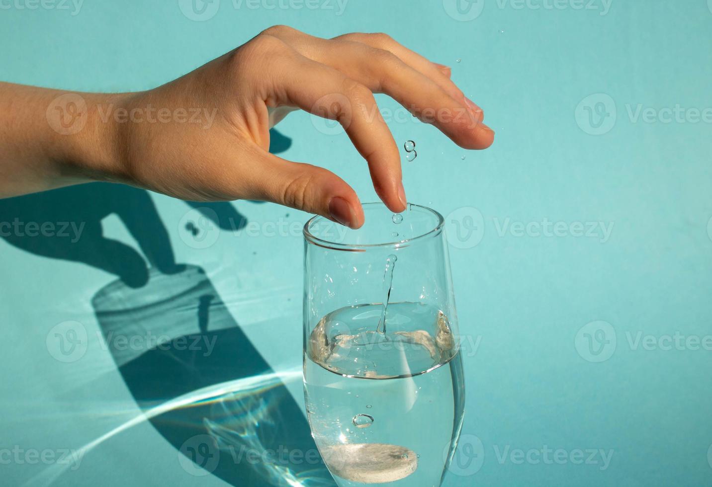 Against a blue background, a hand drops a dissolving fizzy aspirin tablet into a glass of water photo