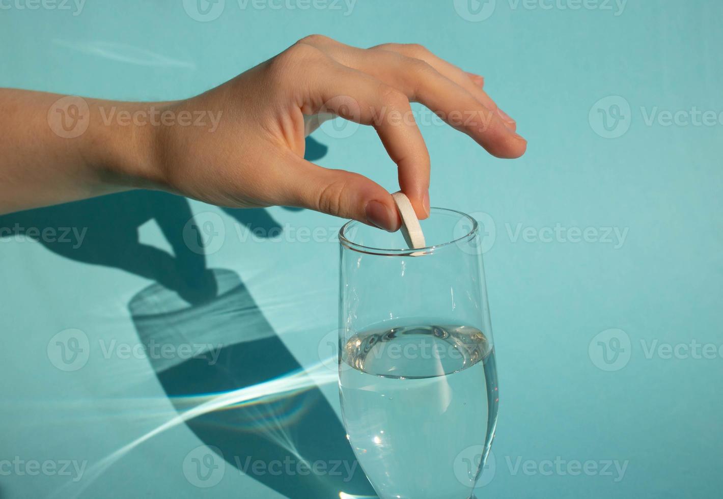 contra un fondo azul, una mano deja caer una tableta de aspirina gaseosa que se disuelve en un vaso de agua foto