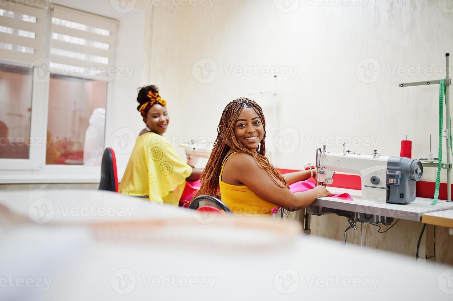 Two african dressmaker woman sews clothes on sewing machine at tailor office. Black seamstress girls. photo
