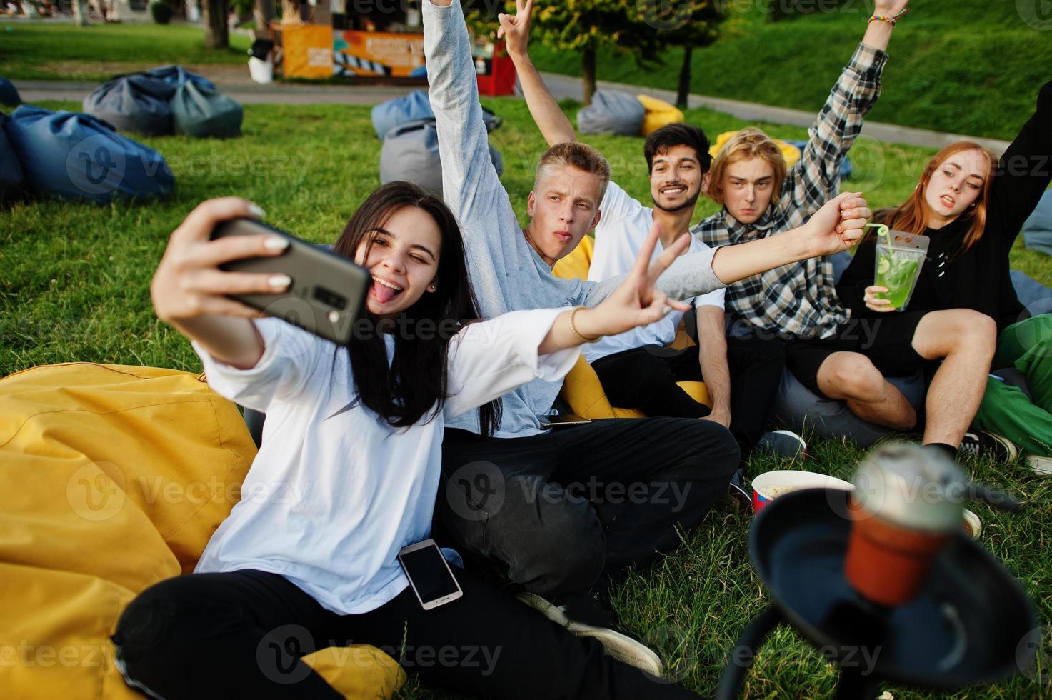 Young multi ethnic group of people watching movie at poof in open air cinema and making selfie on phone. photo