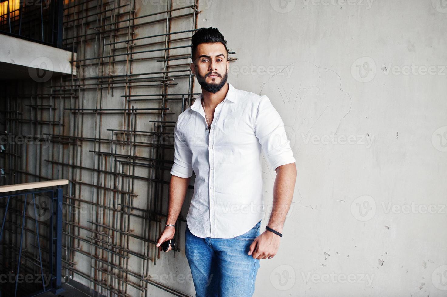 Stylish tall arabian man model in white shirt, jeans posed against steel wall indoor. Beard attractive arab guy. photo