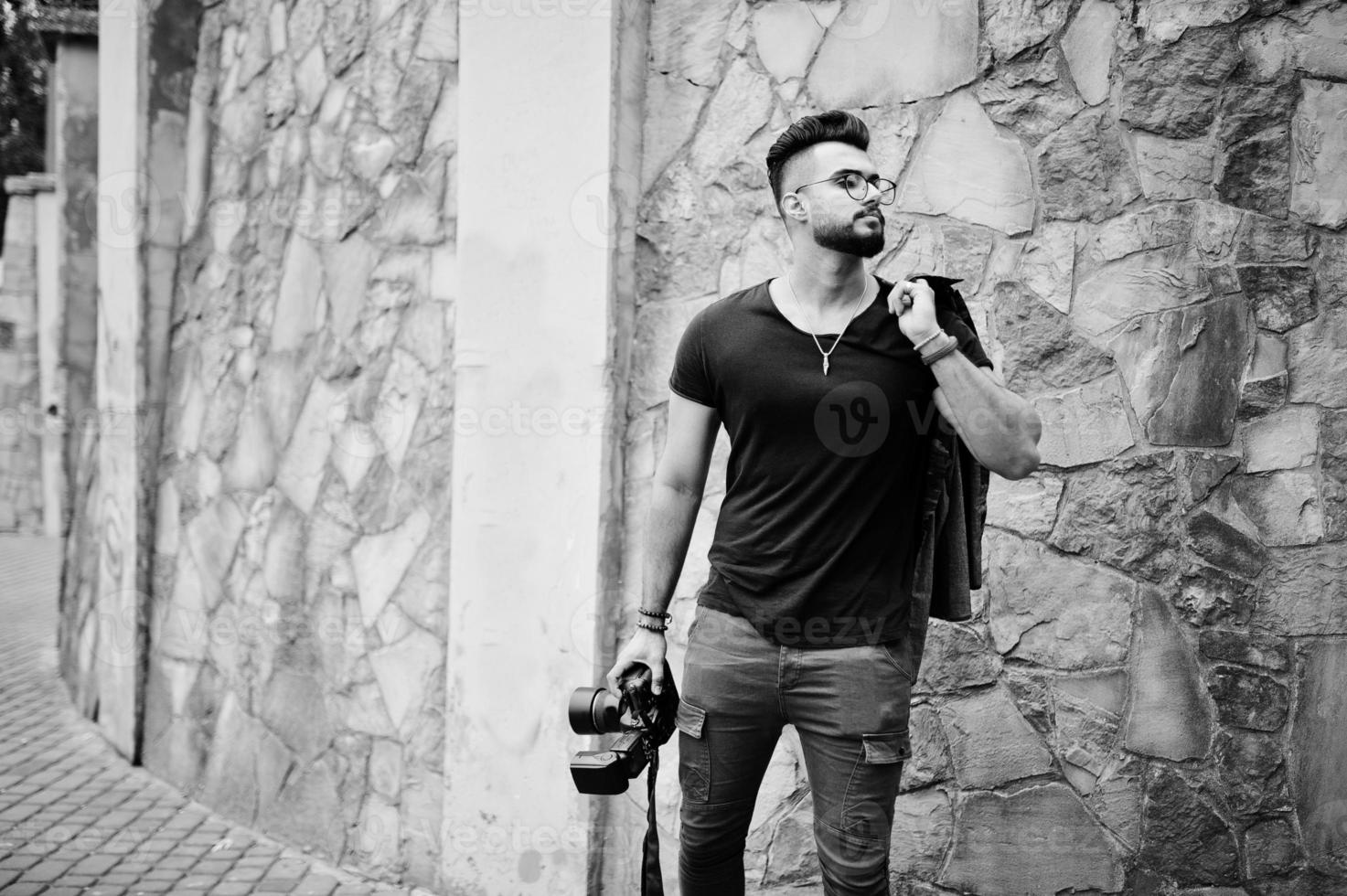 Impresionante y hermoso fotógrafo de hombre macho con barba árabe alta con gafas y camiseta negra con cámara profesional en las manos. foto