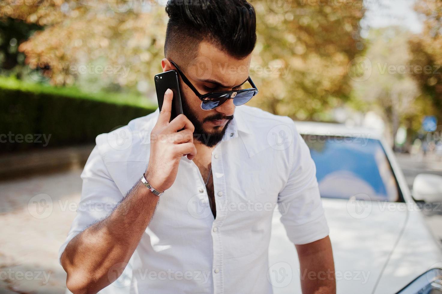 Stylish tall arabian man model in white shirt, jeans and sunglasses posed at street of city. Beard rich attractive arab guy against white suv car, speaking on mobile phone. photo