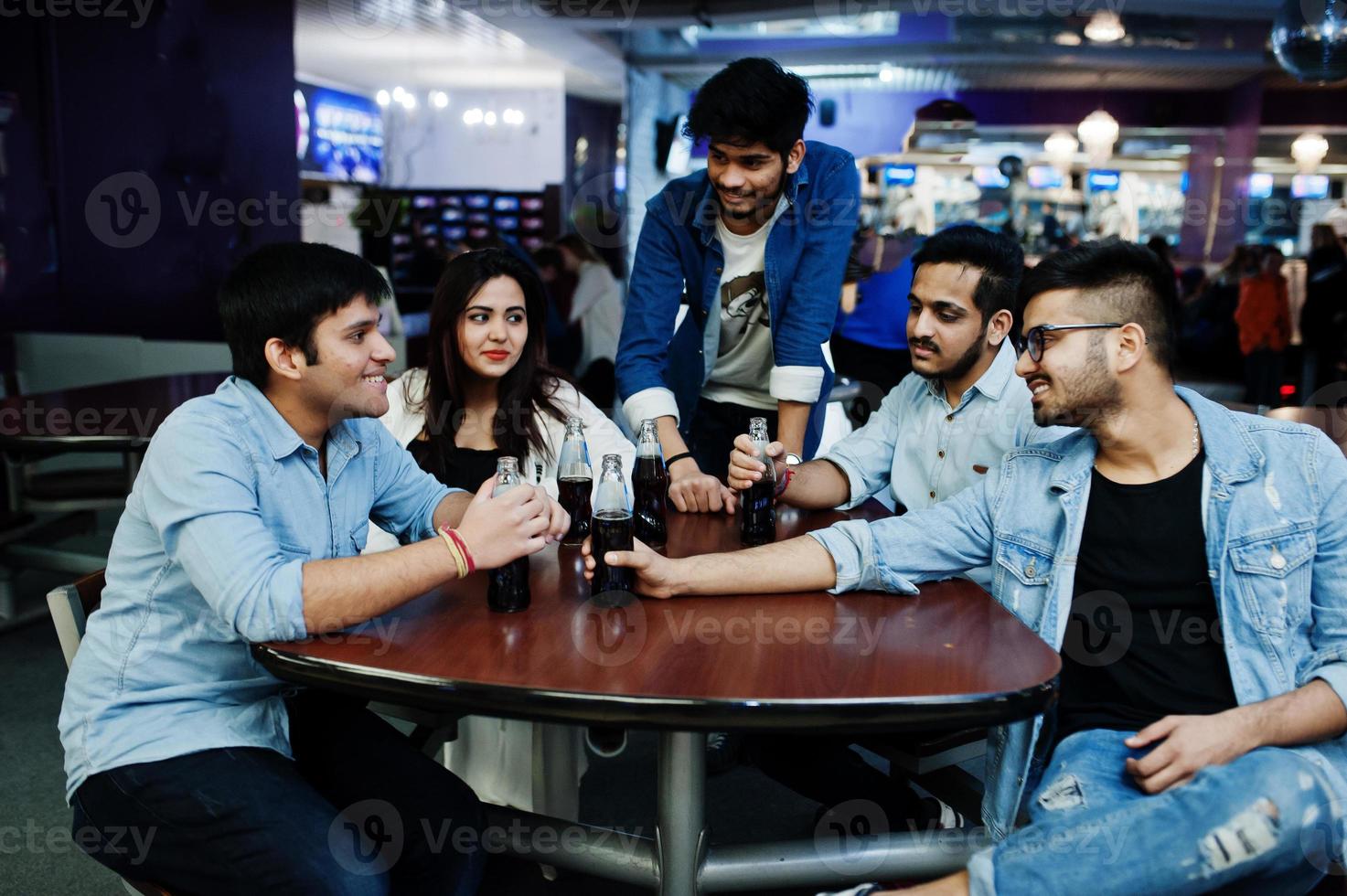 Group of stylish asian friends wear on jeans sitting at table and drink soda from bottles in club. photo