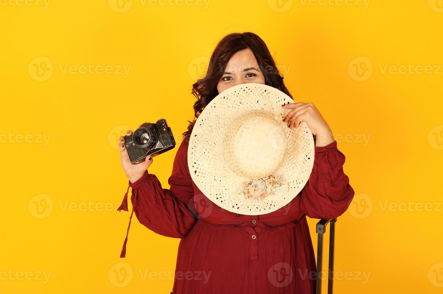 atractiva mujer viajera del sur de asia con vestido rojo intenso, sombrero posado en el estudio sobre fondo amarillo con maleta y cámara de fotos antigua.