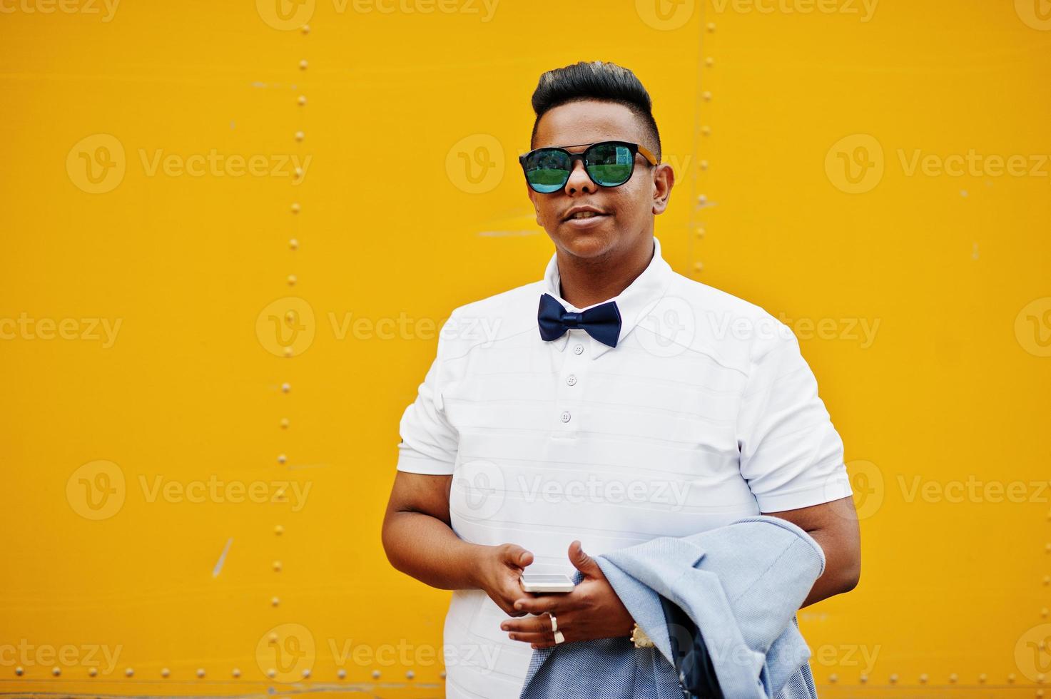 Close up portrait of stylish arabian man at sunglasses and bow tie posed against yellow background with mobile phone. Arab model boy. photo