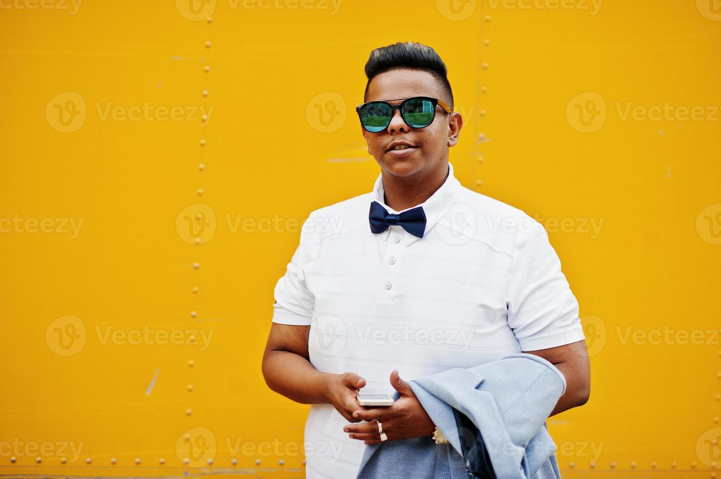 primer plano retrato de un elegante hombre árabe con gafas de sol y corbata de moño posado sobre fondo amarillo con teléfono móvil. chico modelo árabe. foto