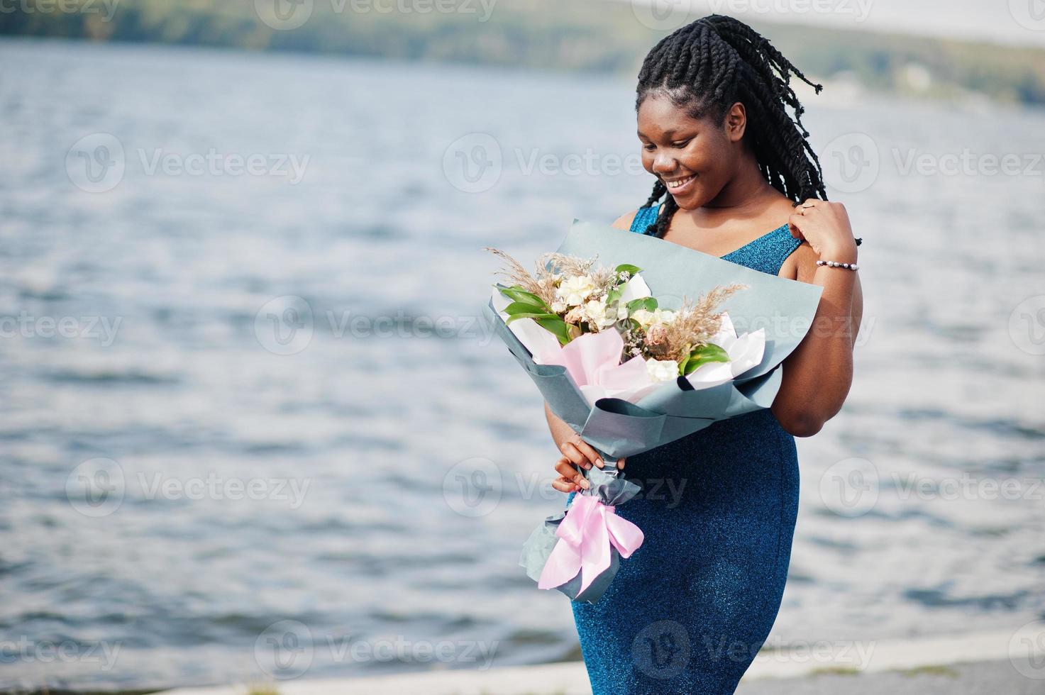 African american dark skinned plus size model posed in a blue shiny dress with bouquet of flowers against sea side. photo