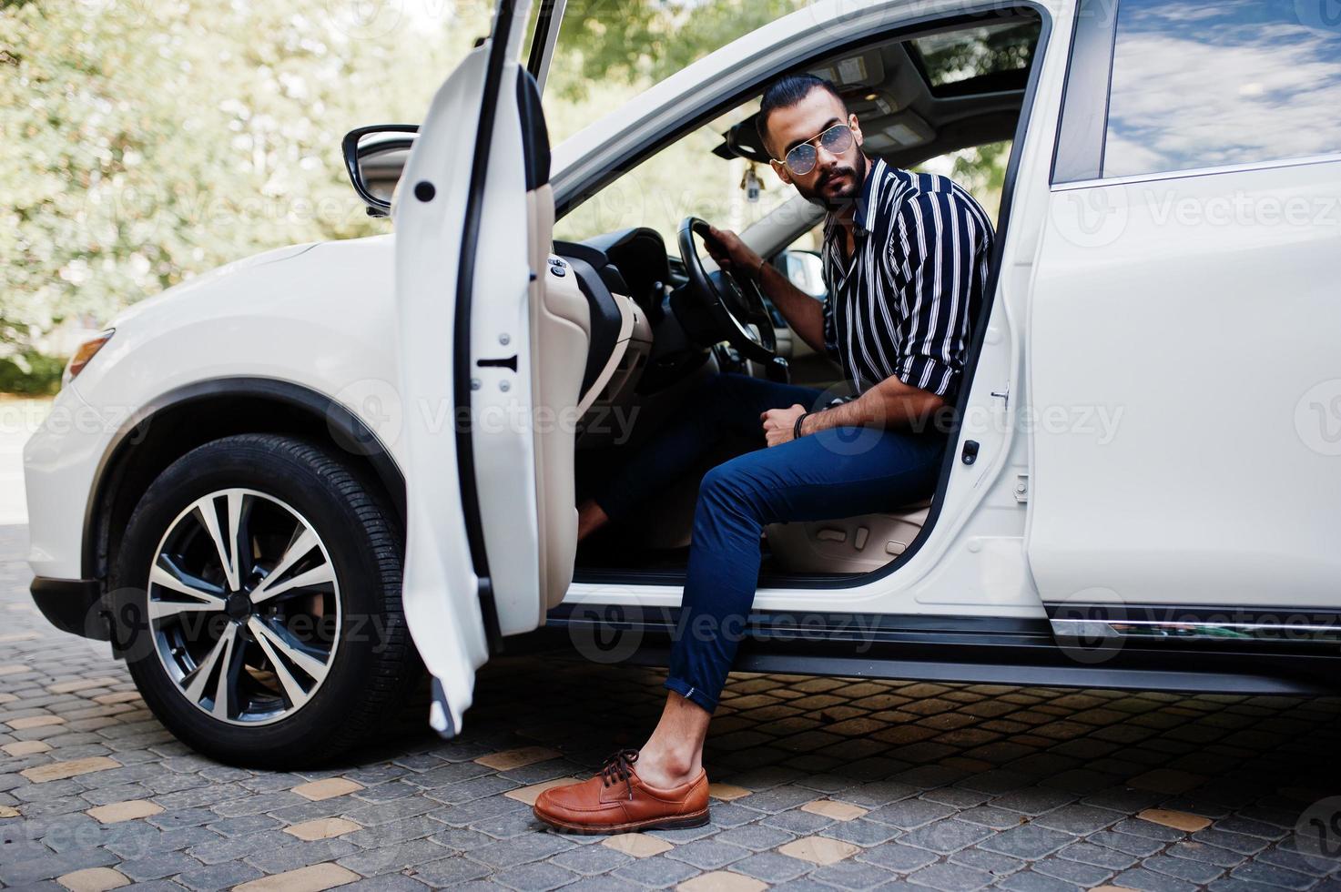 Successful arab man wear in striped shirt and sunglasses pose behind the wheel of  his white suv car. Stylish arabian men in transport. photo